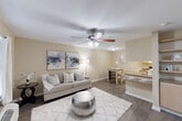 Located at 7575 Willow Chase Boulevard in Houston, Texas, this one-bedroom, one-bathroom unit offers a practical and well-organized single-floor layout. The open-concept living and kitchen area forms the heart of the space, where warm beige walls and durable hardwood-style flooring create a cohesive, neutral aesthetic. A ceiling fan anchors the living room, which receives natural light through a window dressed with grommet-panel curtains. The kitchen is fully equipped with light wood cabinetry, granite-toned countertops, a full suite of white appliances including a range and refrigerator, and a peninsula breakfast bar that provides both additional prep space and casual seating. Tile flooring in the kitchen complements the overall design. The bedroom is a comfortable, well-proportioned retreat featuring its own ceiling fan, a window with blinds and curtains, and direct access to a dedicated walk-in closet with built-in shelving. The bathroom is designed with accessibility in mind, featuring a walk-in shower equipped with grab bars and a fold-down teak bench. Throughout the unit, consistent warm-toned walls and hardwood-style flooring tie the spaces together into a cohesive and functional living environment.