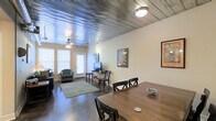 This unique single-floor space makes an immediate impression with its standout corrugated metal ceiling, which lends an industrial yet warm aesthetic throughout the open-plan interior. Dark hardwood floors run continuously underfoot, grounding the space and providing a rich contrast to the light-toned walls.

The open layout seamlessly connects the dining and living areas, creating a cohesive flow ideal for both everyday living and entertaining. The dining area features a solid wood table with ample seating, positioned to take full advantage of the open sightlines across the space. The living area is anchored by a comfortable seating arrangement set atop a soft area rug, with a media setup nearby for relaxed viewing.

Natural light enters generously through multiple windows fitted with white blinds, brightening the interior while a ceiling fan with a light fixture ensures comfortable airflow. Track lighting along the ceiling provides additional illumination and highlights the architectural character of the metal overhead detail.

Wall art adds personality and a creative touch throughout, complementing the space's eclectic, loft-inspired character. Overall, the space offers a well-balanced blend of industrial design elements and comfortable, functional living — making it a visually engaging and practical environment.