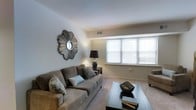 This single-floor space offers a thoughtfully designed layout centered around a comfortable and stylish living area. The living room is bathed in natural light streaming through a wide, multi-pane window dressed with clean white blinds, creating a warm and welcoming atmosphere. Soft wall-to-wall carpeting underfoot adds a cozy feel to the space, while neutral-toned walls provide a versatile backdrop for a range of interior styles. A plush taupe sofa adorned with decorative pillows serves as the focal point of the seating area, paired with a sleek glass-top coffee table that adds a modern touch. A decorative sunburst mirror mounted on the wall introduces an eye-catching artistic element, complemented by a classic table lamp that provides warm ambient lighting. A side table with additional lamp placement enhances the room's functionality and layered lighting. The ceiling features a subtle architectural detail with a recessed ledge, adding visual interest to the otherwise clean and open overhead space. The overall ambiance is calm, polished, and move-in ready, making this a comfortable and functional living environment suited for everyday relaxation.