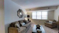 This single-floor space offers a thoughtfully designed layout centered around a comfortable and stylish living area. The living room is bathed in natural light streaming through a wide, multi-pane window dressed with clean white blinds, creating a warm and welcoming atmosphere. Soft wall-to-wall carpeting underfoot adds a cozy feel to the space, while neutral-toned walls provide a versatile backdrop for a range of interior styles. A plush taupe sofa adorned with decorative pillows serves as the focal point of the seating area, paired with a sleek glass-top coffee table that adds a modern touch. A decorative sunburst mirror mounted on the wall introduces an eye-catching artistic element, complemented by a classic table lamp that provides warm ambient lighting. A side table with additional lamp placement enhances the room's functionality and layered lighting. The ceiling features a subtle architectural detail with a recessed ledge, adding visual interest to the otherwise clean and open overhead space. The overall ambiance is calm, polished, and move-in ready, making this a comfortable and functional living environment suited for everyday relaxation.