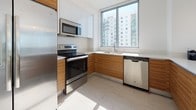 Suite 1601 is a thoughtfully designed high-rise residence that combines contemporary aesthetics with functional living spaces across a single floor. The unit is characterized by an abundance of natural light, delivered through expansive floor-to-ceiling windows and sliding glass doors that frame wide-open city skyline views, giving the space a bright, airy quality throughout.

The open-concept living and dining area flows seamlessly into the kitchen, which features a generous breakfast bar with seating, a crisp white upper cabinetry, warm wood-toned lower cabinets, light tile flooring, and a full suite of stainless steel appliances including a refrigerator, range, microwave, and dishwasher. Pendant lighting accents the bar area, adding a polished finishing touch.

The primary bedroom is serene and carpeted, offering direct access to a private balcony with panoramic views. It connects to a clean, modern en-suite bathroom outfitted with a glass-enclosed shower, floating wood vanity, and large mirror. A second bedroom, also carpeted and well-lit, enjoys its own balcony access and city views. A separate full bathroom with similar modern finishes serves the secondary spaces. The living area balcony is styled with curated décor, lush tropical greenery, and a geometric gold mirror, enhancing the elevated urban atmosphere of the suite.