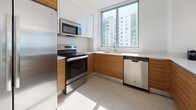 Suite 1601 is a thoughtfully designed high-rise residence that combines contemporary aesthetics with functional living spaces across a single floor. The unit is characterized by an abundance of natural light, delivered through expansive floor-to-ceiling windows and sliding glass doors that frame wide-open city skyline views, giving the space a bright, airy quality throughout.

The open-concept living and dining area flows seamlessly into the kitchen, which features a generous breakfast bar with seating, a crisp white upper cabinetry, warm wood-toned lower cabinets, light tile flooring, and a full suite of stainless steel appliances including a refrigerator, range, microwave, and dishwasher. Pendant lighting accents the bar area, adding a polished finishing touch.

The primary bedroom is serene and carpeted, offering direct access to a private balcony with panoramic views. It connects to a clean, modern en-suite bathroom outfitted with a glass-enclosed shower, floating wood vanity, and large mirror. A second bedroom, also carpeted and well-lit, enjoys its own balcony access and city views. A separate full bathroom with similar modern finishes serves the secondary spaces. The living area balcony is styled with curated décor, lush tropical greenery, and a geometric gold mirror, enhancing the elevated urban atmosphere of the suite.