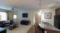 This single-floor residence showcases an open-concept design that integrates the living, dining, and kitchen areas into one cohesive space. The living area features plush carpeting, a ceiling fan, and a wall-mounted flat-screen television above a dark wood media console. Natural light filters in through a window dressed with blinds, contributing to the room's bright, neutral ambiance. Crown molding accents the white ceilings, adding a refined architectural touch throughout.

The kitchen and dining area transition naturally from the living space, marked by warm hardwood-style flooring and a raised bar counter topped with dark granite. Pendant lighting hangs above the bar, providing focused illumination over the seating area, which features patterned upholstered chairs. The kitchen includes a stainless steel sink and dark cabinetry, maintaining a consistent, sophisticated color palette.

An arched hallway leads to additional rooms beyond, adding architectural character while connecting the living spaces. The overall aesthetic is cohesive, combining neutral tones, dark wood accents, and contemporary fixtures to create a comfortable and well-appointed interior.
