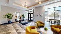 This thoughtfully designed amenity floor offers a seamless blend of elegance and functionality across a generous single-level layout. The entrance lobby immediately impresses with its striking geometric marble-look tile flooring, a coffered ceiling adorned with a sculptural sputnik chandelier, and floor-to-ceiling glass doors that flood the space with natural light and urban views. Plush gold velvet armchairs and a console table with curated decor create an inviting lounge atmosphere, while a wet bar with teal cabinetry and dark countertops adds a refined, hospitality-inspired touch.

The hallway extends from the lobby with a clean, modern aesthetic, featuring patterned carpeting, dark-framed numbered doors with gold accents, and warm wall sconces. A dedicated mailroom area is anchored by a rich wood accent wall housing resident mailboxes, lit by globe pendant chandeliers overhead.

The fitness center is a standout space, boasting warm hardwood floors, floor-to-ceiling windows offering sweeping outdoor views, bold decorative wallpaper, wall-to-wall mirrors, and modern pendant lighting. Cardio equipment is well-positioned to take advantage of the natural light. Restroom facilities are also accessible from the lobby corridor, finished with dark panel doors and elegant signage, completing this polished and well-appointed amenity level.