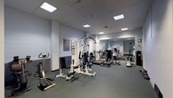 The fitness center is a functional, single-floor exercise facility designed to accommodate a range of workout routines. The space features a clean, utilitarian aesthetic with light gray walls and a drop ceiling fitted with recessed fluorescent panels that provide even, bright illumination throughout. The flooring consists of durable green exercise matting, offering comfort and stability during workouts. A large wall-mounted mirror spans a significant portion of one side of the room, visually expanding the space and allowing users to monitor their form. The equipment selection includes a multi-station cable and weight machine suitable for a variety of strength training exercises, along with treadmills for cardiovascular conditioning and a stationary bike for low-impact aerobic workouts. Informational placards and instructional guides are posted on the walls, offering guidance on proper equipment use. The open layout ensures adequate space between machines for safe movement and circulation. Overall, the fitness center presents a straightforward and practical environment well-suited for daily exercise needs.