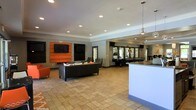 This single-story communal space is designed as a multifunctional lounge and entertainment area. The open-plan layout is anchored by a bold orange media wall, which houses a flat-screen television and framed display panels, creating a vibrant focal point against the neutral grey walls. Recessed lighting throughout the ceiling provides consistent, even illumination across the space.

The lounge area features a large, dark leather sectional sofa complemented by a charcoal accent chair, offering generous seating for residents or guests. An orange armchair near the perimeter adds a cohesive pop of color. The flooring consists of large-format stone-look tiles, contributing to a clean, contemporary aesthetic.

Beyond the seating area, the space opens into a dedicated game room, visually separated by a structural column. This zone includes what appears to be a shuffleboard or billiards table, along with additional seating along the walls. A sleek white counter or bar-height surface is positioned near the transition between zones, potentially serving as a refreshment or reception area. A pendant light fixture adds a refined touch overhead, completing the modern, amenity-rich character of the space.