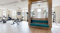 Located at Sawgrass Bay in Clermont, FL, this fitness center offers a spacious, single-floor layout designed to support a full range of workout routines. The space features light hardwood-style flooring, coffered ceilings with recessed lighting, and multiple ceiling fans that contribute to a bright and comfortable environment. Large windows line the walls on multiple sides, flooding the interior with natural light and providing views of the surrounding community. The main workout area is well-appointed with a variety of cardio equipment, including treadmills, ellipticals, a stationary bike, and a rowing machine, as well as wall-mounted TVs for entertainment during workouts. A dedicated strength training section houses a cable machine, a power rack with an adjustable bench, a dumbbell rack, and additional free weights. Full-length mirrors along one wall enhance the sense of openness and allow for form-checking during exercise. A distinctive built-in wooden nook with teal upholstered seating and integrated shelving serves as a lounge or stretching area, adding a polished, inviting touch to the space. Contemporary abstract artwork is displayed throughout, and glass-panel entry doors provide a clean, modern aesthetic. The overall ambiance is airy, modern, and functional.