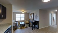 This thoughtfully designed residence at Jefferson Crossings Senior Living offers a comfortable and functional living environment tailored to the needs of its residents. The single-floor layout ensures ease of movement throughout the space, making daily life both convenient and accessible.

The living area serves as the heart of the home, featuring warm hardwood-style flooring that adds a clean, modern aesthetic. A ceiling fan with integrated lighting keeps the space comfortable year-round, while a large window invites generous natural light and provides pleasant outdoor views. The open feel of the room is enhanced by neutral wall tones and a well-defined layout that comfortably accommodates a full seating arrangement and a dining area side by side.

The dining space is neatly integrated into the living area, featuring a compact table with seating — ideal for casual meals or social gatherings. The overall design strikes a balance between practicality and warmth, with decorative wall art adding personality to the space. The cohesive color palette of soft grays, tans, and cheerful yellow accents gives the residence a welcoming and uplifting atmosphere. Every detail of this home reflects a commitment to comfortable, independent senior living within a well-appointed community setting.