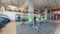 Located at 1600 W Lincoln Ave in Anaheim, California, this two-story fitness facility offers a comprehensive and thoughtfully designed workout environment across a generous total footprint. The larger main floor serves as the heart of the facility, featuring soaring ceilings accented by a striking grid-style pendant lighting system and bold structural columns. This expansive space is divided into distinct training zones, including a functional turf training area equipped with sleds and tire equipment, a strength zone with color-coded Olympic barbells and squat racks, and a machine circuit section. Large arched windows flood the floor with natural light, enhancing the open and energizing atmosphere. A dedicated Pilates studio on this level features warm wood-tone flooring, a sleek wood-paneled ceiling with recessed linear lighting, full-length mirrors, and glass wall partitions that provide both visibility and privacy. The upper floor offers a well-appointed cardio mezzanine with wood flooring, treadmills, stationary bikes, and stair climbers positioned near floor-to-ceiling windows overlooking the exterior. A separate exercise room on the upper level features distinctive arched windows with warm wood accents and an open layout ideal for cycling and functional training. The facility's modern industrial aesthetic, dual-floor layout, and diverse equipment selection make it a complete and high-caliber fitness destination.