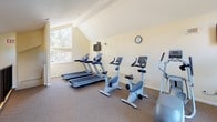 This two-story fitness center at 5253 El Cerrito Dr in Riverside, California, offers a well-equipped workout facility designed to support a full range of exercise routines. The space features a vaulted, sloped ceiling that lends an open, airy feel, complemented by large windows on both floors that flood the interior with natural light and provide views of the surrounding mature trees and palm trees outdoors.

The lower level houses a dedicated strength training area, anchored by a multi-function cable machine ideal for a variety of resistance exercises. A staircase connects the two floors, with a black metal railing adding a clean, modern detail to the interior. The upper level is devoted to cardiovascular training and features multiple treadmills, upright stationary bikes, and elliptical machines arranged to take full advantage of the natural light streaming in from the surrounding windows. A wall-mounted television is positioned for easy viewing during workouts.

Throughout both levels, neutral-toned carpet provides comfortable footing, while the warm cream-colored walls contribute to a bright and welcoming atmosphere. Direct exterior door access on the upper level offers convenient entry and exit. The overall layout is functional and spacious, making this fitness center a practical and well-appointed amenity.