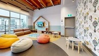 Located at 1915 Sooke Rd in Colwood, BC, this thoughtfully designed children's amenity room delivers a lively and welcoming environment for young residents. The space is defined by its striking exposed wood-beam ceiling, which adds warmth and architectural character, complemented by sleek linear pendant lighting suspended throughout.

A large floor-to-ceiling window wall floods the interior with natural light and offers views of the surrounding neighbourhood. The open layout is cleverly zoned to serve different age groups and activities. A central circular play area with a polished concrete floor anchors the room, surrounded by colorful bean bag chairs in yellow, orange, and white. Adjacent to this is a raised play nook framed by a charming house-shaped wood surround, complete with built-in storage shelving, a wall-mounted TV, and a soft play mat — ideal for younger children.

A dedicated activity corner features child-sized tables and chairs alongside a whimsical hot air balloon-patterned accent wall that adds a sense of imagination and adventure. A modular seating area in soft pastel tones — cream, sage, and orange — sits beneath the large windows, offering a comfortable spot for caregivers. A small service counter with a sink provides practical functionality. A chalkboard panel and access to an adjacent fitness room round out this well-appointed, family-friendly space.