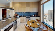 This single-floor residence showcases a thoughtfully curated interior design that seamlessly blends natural materials with contemporary elegance throughout its well-appointed rooms. The kitchen features light blonde wood cabinetry extending to the ceiling, dark stone countertops, a dramatic slate-toned backsplash, stainless steel appliances including a built-in wine refrigerator, and a rustic wood dining table surrounded by leather chairs. The dining room centers on a striking marble-topped table beneath a sculptural chandelier, accompanied by bold abstract artwork and grasscloth-textured walls. The living area offers light hardwood flooring, textured wall treatments, and floor-to-ceiling windows framing expansive panoramic city and mountain views. A dedicated study features a distinctive organically shaped solid wood desk, sisal-tone carpeting, and generous corner windows capturing sweeping cityscape views. The primary bedroom is serene and spacious, with a large picture window overlooking the city skyline, a sculptural ceiling light fixture, and hardwood floors. Two bathrooms are finished with natural stone tile flooring; one includes a freestanding soaking tub and a clean-lined floating vanity, while the other features mosaic tile walls and a walk-in shower. Additional highlights include a well-organized walk-in closet with custom warm wood shelving and a secondary bedroom with built-in cabinetry.