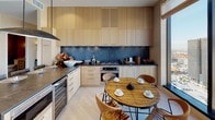This single-floor residence showcases a thoughtfully curated interior design that seamlessly blends natural materials with contemporary elegance throughout its well-appointed rooms. The kitchen features light blonde wood cabinetry extending to the ceiling, dark stone countertops, a dramatic slate-toned backsplash, stainless steel appliances including a built-in wine refrigerator, and a rustic wood dining table surrounded by leather chairs. The dining room centers on a striking marble-topped table beneath a sculptural chandelier, accompanied by bold abstract artwork and grasscloth-textured walls. The living area offers light hardwood flooring, textured wall treatments, and floor-to-ceiling windows framing expansive panoramic city and mountain views. A dedicated study features a distinctive organically shaped solid wood desk, sisal-tone carpeting, and generous corner windows capturing sweeping cityscape views. The primary bedroom is serene and spacious, with a large picture window overlooking the city skyline, a sculptural ceiling light fixture, and hardwood floors. Two bathrooms are finished with natural stone tile flooring; one includes a freestanding soaking tub and a clean-lined floating vanity, while the other features mosaic tile walls and a walk-in shower. Additional highlights include a well-organized walk-in closet with custom warm wood shelving and a secondary bedroom with built-in cabinetry.