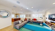 This thoughtfully designed single-floor space serves as a versatile communal hub, offering a range of functional areas that flow seamlessly into one another. The open-plan lounge and kitchen area forms the heart of the space, where a generously sized sectional sofa in warm gray anchors a vibrant teal and blue striped area rug. Colorful accent pillows and eclectic wall art add personality, while recessed lighting and multiple windows allow natural light to fill the room. A large flat-screen TV and a classic plaid accent chair complete the media lounge area. Adjacent to the lounge, the kitchen features a substantial island with bar seating, warm wood cabinetry, stainless steel appliances, and a striking geometric pendant light that serves as a focal centerpiece. A dedicated dining room accommodates group gatherings with multiple tables and cross-back chairs set on light hardwood floors, with large windows providing a bright, airy atmosphere. A quiet business center offers a clean workspace with a computer setup, ergonomic seating, and a tall indoor plant for a refreshing touch. The restroom is finished with charming hexagonal teal and cream floor tiles and includes accessibility-friendly fixtures. Curated framed artwork is displayed throughout, adding a cohesive, polished aesthetic across the entire space.
