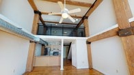 This two-floor loft-style space masterfully blends industrial character with modern functionality. The main level opens into a dramatic, double-height living area defined by exposed heavy timber beams with iron bracket accents, a structural column, and warm hardwood flooring throughout. A ceiling fan anchors the soaring ceiling, while exposed ductwork adds to the industrial aesthetic. The open-concept kitchen is positioned along one wall and features light maple cabinetry, a dark countertop with an integrated sink, stainless steel appliances, and pendant lighting overhead. The kitchen flows seamlessly into the main living area, creating a cohesive and versatile space. Original exposed brick is visible along the perimeter, lending warmth and historic character to the interior. A carpeted staircase with a sleek black metal railing leads to the upper mezzanine level, which overlooks the main floor through a glass and metal railing system, maintaining an open, airy feel between levels. Windows allow natural light to filter into the space, enhancing the overall brightness. The combination of raw industrial elements — brick, timber, and metal — alongside contemporary finishes creates a distinctive and richly textured environment well-suited for both living and creative use.