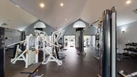 This fitness center is housed in a striking single-story structure defined by dramatic vaulted ceilings that rise to impressive heights, creating an open and energizing workout environment. Distinctive arched windows near the ceiling peaks allow natural light to pour in, complementing the recessed lighting throughout and keeping the space bright and welcoming.

The facility is divided into dedicated zones for both strength training and cardiovascular exercise. The strength area is well-stocked with a comprehensive selection of HOIST cable machines, multi-function weight stations, adjustable benches, and a full dumbbell rack ranging across multiple weight increments. Large wall-mounted mirrors visually expand the space and allow users to monitor their form during workouts.

The cardio zone is positioned near floor-to-ceiling glass windows and sliding doors that offer direct views of an outdoor pool and lush tree-lined surroundings. This area features treadmills, elliptical trainers, and stationary bikes from leading fitness brands, with a wall-mounted television nearby for entertainment.

Dark rubberized flooring runs throughout, providing durability and safety underfoot. The walls are finished in a cool gray tone, contributing to a focused and modern aesthetic. Access to the outdoor area is available through both glass-paned double doors and large sliding glass panels, seamlessly connecting the interior fitness space to the exterior amenities.