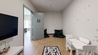 Located at 1819 Avenue Dollard in Montréal, this thoughtfully designed amenity space serves as a party room and playground for residents. The interior is bright and contemporary, featuring light hardwood flooring, recessed lighting, and a neutral palette that creates a welcoming atmosphere throughout.

The kitchen is outfitted with light wood cabinetry, white upper cabinets, a stainless steel range, integrated refrigerator, and a matte black faucet over a single-basin sink — combining functionality with a clean, modern aesthetic.

The dining area is a standout space, anchored by a large dark wood table surrounded by curved, upholstered chairs. Floor-to-ceiling windows flood the room with natural light and offer direct views of the outdoor playground. An exposed concrete structural column adds an industrial design element, while a wall-mounted flat-screen TV and a wood sideboard complete the space.

A dedicated children's room labeled "Salle Enfants" features playful polka-dot wallpaper, bean bag chairs, a small table and chairs set, and a play mat — creating an engaging, child-friendly environment.

Outside, a lush artificial turf playground provides ample open space with fitness and play structures, set against the backdrop of the residential building's modern façade with glass-railed balconies.