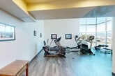 Located at 200 E Tenth Street in Gilroy, California, this fitness center offers a well-appointed, single-room exercise space designed to support a variety of workout routines. The room is characterized by soaring ceilings that lend an open, airy feel, complemented by clean white walls and durable hardwood-style flooring throughout.

A dramatic floor-to-ceiling window wall on one side of the room floods the space with abundant natural light while providing expansive views of the surrounding outdoor environment, creating an energizing atmosphere for exercise. An accent soffit with warm golden tones adds a distinctive architectural detail near the entrance area.

The fitness center is equipped with a comprehensive selection of exercise machines, including multiple elliptical trainers, a treadmill, a cable weight machine, and a rowing machine, along with a flat workout bench — offering options for both cardiovascular and strength training. A small wooden side table provides a convenient surface near the workout area.

Framed photographs and signage referencing local history are displayed along the walls, adding a sense of place and community character to the space. Ample floor space between equipment ensures comfortable movement throughout the room, making this fitness center a functional and welcoming facility for users at all fitness levels.