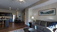 This single-story space offers an open-concept design that seamlessly connects the living room, dining area, and kitchen into one cohesive environment. The living area is anchored by a comfortable gray sofa, a patterned area rug, and a dark coffee table, creating a welcoming focal point. A ceiling fan overhead provides both function and a finishing touch to the space. Neutral taupe and white wall tones carry throughout, contributing to a clean, modern aesthetic, while framed wall art adds a subtle decorative element. Dark hardwood-style flooring runs continuously through the space, grounding the design with warmth and contrast against the lighter walls. A console table with a table lamp and decorative accents provides additional character near the living area. Through an open archway, a compact dining nook is visible, furnished with a bistro-style table and bar stools, conveniently positioned adjacent to the kitchen. The kitchen features stainless steel appliances, including a full-sized refrigerator. A white paneled door leads to additional rooms beyond, suggesting further private living spaces. Overall, the layout is efficient and well-appointed.