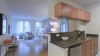 This single-floor residence offers a thoughtfully designed open-concept layout that connects the kitchen and living areas into one cohesive, welcoming space. The kitchen features warm honey-toned wood cabinetry, dark stone countertops, and a peninsula that doubles as a casual dining or prep area, complete with a stainless steel sink. An arched decorative mirror adds an elegant architectural touch to the kitchen's backdrop. The living area is generously proportioned, bathed in natural light streaming through multiple large windows dressed with classic white curtains. Light hardwood flooring runs throughout, adding warmth and continuity to the space. A tasteful area rug anchors the seating arrangement, which includes upholstered armchairs and a sofa styled with accent pillows. Overhead, a decorative pendant light fixture provides ambient illumination and a focal point for the room. Neutral wall tones in soft gray create a calm, sophisticated palette that complements the natural light. The overall ambiance is bright, airy, and elegantly understated, making it a comfortable and functional living environment suited for both everyday living and entertaining.