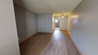 This single-floor unit presents a practical and well-organized layout designed for comfortable everyday living. The main living area is finished with durable wood-look laminate flooring and neutral gray walls, creating a bright and clean atmosphere. A textured ceiling runs throughout the common areas, adding subtle dimension to the space. Natural light enters through an exterior door that opens to an outdoor area, enhancing the openness of the living zone.

The galley-style kitchen runs along one wall and is efficiently designed with white cabinetry both above and below the countertops, offering ample storage. A stainless steel double-basin sink with a matte black faucet anchors the countertop, which features a speckled laminate finish. A white electric range completes the cooking area, and the light wood-look flooring continues seamlessly from the main living space.

The bedroom is carpeted for warmth and comfort, featuring neutral gray walls and a ceiling fan with an integrated light fixture. A window with blinds allows for natural light and ventilation.

The bathroom includes a large vanity mirror above a drop-in sink set into a speckled laminate countertop, with a matte black faucet. A shower-tub combination with a decorative curtain completes the space. Throughout, the unit maintains a cohesive, understated aesthetic.
