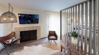 This single-floor residence showcases a cohesive modern design with warm wood-look flooring and a neutral palette carried throughout. The open-concept kitchen is outfitted with gray shaker-style cabinetry, granite-look countertops, and a full suite of stainless steel appliances, including a refrigerator, dishwasher, microwave, and range. A built-in dining counter with bar stools provides a casual yet functional dining area, making efficient use of the space. The living area features a modern tiled fireplace flanked by built-in storage, a wall-mounted TV, and a distinctive vertical slat partition that elegantly separates it from the bedroom zone, allowing natural light to filter through sheer curtains. The bedroom is a serene retreat, highlighted by pendant lighting, recessed ceiling lights, and a striking longhorn canvas artwork above the bed. A sliding barn door adds a contemporary touch and leads to additional areas of the unit. The bathroom is clean and well-finished, featuring gray cabinetry, granite-look countertops, a framed mirror, and wood-look flooring that ties seamlessly to the rest of the space. Every detail reflects a carefully curated, modern aesthetic designed for comfortable single-floor living.