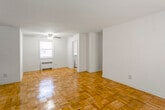 This single-floor residence is defined by its warm, classic aesthetic, most notably the richly patterned parquet wood flooring that runs consistently throughout the living areas and bedrooms, lending the space a timeless character. The open living and dining area is generously proportioned, bathed in natural light from a window overlooking greenery, and equipped with a ceiling fan. A radiator provides in-unit heating. The layout flows naturally through a central hallway connecting the main rooms. The bedroom is well-sized, featuring two windows that allow ample natural light and outdoor views, a ceiling fan, and the same parquet flooring. The kitchen is a separate, dedicated space with light wood-tone flooring, white cabinetry, a stainless steel sink with countertop, and a white gas range, with a window providing natural light. The bathroom is finished in white ceramic tile on both walls and floors, featuring a single-basin vanity with wood cabinetry, a bathtub with shower, a radiator, and a frosted privacy window. Throughout the unit, white walls and clean lines create a bright, neutral backdrop.