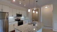 The kitchen serves as the heart of this single-floor space, showcasing a thoughtfully designed layout that balances functionality and style. White shaker-style cabinetry lines the walls, offering generous storage while maintaining a clean, bright aesthetic. The countertops throughout are finished in light granite with natural veining, adding an elegant, timeless quality to the space.

A central island anchors the kitchen, featuring an undermount stainless steel sink with a modern gooseneck faucet. Three pendant lights hang above the island in varying styles, providing focused task lighting as well as a decorative focal point. Additional track lighting installed along the ceiling ensures the workspace is well-illuminated throughout.

Stainless steel appliances, including a built-in microwave and a freestanding range with an electric cooktop, complement the cabinetry seamlessly. A classic subway tile backsplash adds texture behind the range area. Crown molding runs along the ceiling perimeter, lending an elevated, finished look to the space. A hallway visible beyond the kitchen hints at further rooms, connected by arched doorways that contribute to the overall architectural character.
