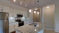 The kitchen serves as the heart of this single-floor space, showcasing a thoughtfully designed layout that balances functionality and style. White shaker-style cabinetry lines the walls, offering generous storage while maintaining a clean, bright aesthetic. The countertops throughout are finished in light granite with natural veining, adding an elegant, timeless quality to the space.

A central island anchors the kitchen, featuring an undermount stainless steel sink with a modern gooseneck faucet. Three pendant lights hang above the island in varying styles, providing focused task lighting as well as a decorative focal point. Additional track lighting installed along the ceiling ensures the workspace is well-illuminated throughout.

Stainless steel appliances, including a built-in microwave and a freestanding range with an electric cooktop, complement the cabinetry seamlessly. A classic subway tile backsplash adds texture behind the range area. Crown molding runs along the ceiling perimeter, lending an elevated, finished look to the space. A hallway visible beyond the kitchen hints at further rooms, connected by arched doorways that contribute to the overall architectural character.
