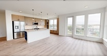 This contemporary single-floor residence offers 1,180 square feet of thoughtfully designed living space across 2 bedrooms and 2 bathrooms. Light-toned hardwood flooring flows throughout, complementing the unit's clean, modern aesthetic defined by white walls and black hardware accents.

The open-concept kitchen and living area serves as the heart of the home. The kitchen features warm wood-grain cabinetry, white quartz countertops, stainless steel appliances, a large center island with pendant lighting, open shelving, and a built-in window bench with storage — all bathed in natural light. The living area is lined with a row of large windows and a glass balcony door, providing sweeping outdoor views and direct balcony access, with a ceiling fan overhead.

The primary bedroom measures 150 square feet and includes a ceiling fan and generous windows with exterior views. It connects to a well-appointed en-suite bathroom featuring a dual-sink vanity with a bold blue cabinet, backlit mirror, glass-enclosed walk-in tile shower, and direct access to a walk-in closet with built-in shelving and hanging rods. The second bedroom also features a ceiling fan, bright windows with views, and its own closet. Additional spaces include a dedicated laundry room and two hallways connecting the layout efficiently.