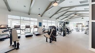 This generously sized fitness center occupies a single floor and is designed to accommodate a wide range of workout needs. The architectural centerpiece is the vaulted ceiling adorned with exposed wood beams, lending a modern farmhouse aesthetic to the space. Pendant light fixtures hang throughout, complementing the abundant natural light that streams in through large floor-to-ceiling windows lining multiple walls. These windows also provide open outdoor views, creating an airy and inviting atmosphere.

The fitness floor is divided into distinct functional zones. The cardio area is lined with treadmills, ellipticals, stationary bikes, and rowing machines, all positioned near the expansive windows. A dedicated strength training zone features a power rack, benches, a dumbbell rack, and multiple cable and resistance machines. A separate functional training area includes a heavy bag, battle ropes, medicine balls, resistance bands, and a multi-purpose rig, offering space for dynamic and high-intensity workouts.

A bold geometric mural in dark gray and white anchors one interior wall, adding a striking visual element. The neutral-toned flooring ties the space together, while direct exterior access doors provide convenient entry and exit throughout the facility.