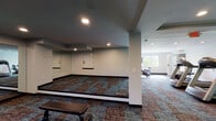 Located at 3040 Suntree Plaza in Kansas City, Kansas, this fitness center offers a well-organized, single-floor layout designed to accommodate a variety of workout routines. The space is divided into distinct functional zones, each tailored to a specific type of exercise.

The cardio area is equipped with Precor treadmills, an elliptical trainer, and a stationary bike, all positioned near windows that allow natural light to fill the room and provide views of the surrounding greenery. A wall-mounted flat-screen television adds convenience for users during cardio sessions.

Adjacent to the cardio zone is a free weights area, featuring a multi-tier dumbbell rack stocked with a range of weights alongside a padded workout bench. Large floor-to-ceiling mirrors line the walls in this section, enhancing the sense of space and allowing users to monitor their form during exercises.

A dedicated open-floor stretching and exercise area completes the layout, offering ample room for mat work, mobility exercises, or bodyweight training. This section also features mirrors along the wall and a low platform area.

Throughout the facility, patterned carpet flooring in earthy tones of blue, brown, and rust adds a modern aesthetic. Recessed lighting illuminates the entire space uniformly, complementing the natural light from multiple windows. The overall design is clean, functional, and well-suited for a comprehensive fitness routine.