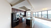 Located in the heart of downtown Cincinnati on E 6th Street, this high-rise unit offers a compelling blend of modern finishes and dramatic urban views. The open-concept living and kitchen area is anchored by a large granite-topped island with a stainless steel sink and pendant lighting, seamlessly connecting the cooking and living spaces. The kitchen is fully equipped with rich dark-cherry cabinetry, a tile backsplash, granite countertops, and a full suite of stainless steel appliances. Wood-look tile flooring flows throughout the main living area, adding warmth and continuity. The living space is defined by a stunning wall of floor-to-ceiling corner windows that bathe the interior in natural light and frame sweeping panoramic views of the Cincinnati skyline and surrounding hillsides. The bedrooms feature plush carpeting, ceiling fans, and large windows that continue the city view experience. Two bathrooms are appointed with dark-wood vanities, white countertops, framed mirrors, and large-format tile flooring. The primary bathroom includes an adjoining walk-in closet. The overall design balances contemporary urban aesthetics with functional, well-finished interiors suited for elevated city living.