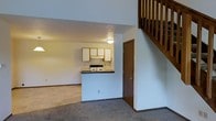 This two-story space features an open-concept main level where the living area and kitchen are seamlessly connected. The living area is finished with wall-to-wall carpeting, creating a comfortable and cohesive feel throughout the lower floor. The kitchen is positioned at the far end of the main level, defined by a peninsula-style countertop with dark laminate surfaces that separates it from the living space. White cabinetry lines the kitchen walls, complemented by a range with overhead ventilation. The kitchen's layout is compact yet functional, with a door nearby providing additional access. A wooden staircase with a classic balustrade rises along one wall, leading to the upper floor, which overlooks the main level below through a railing. The soaring vertical space created by the two-story layout adds an airy, open quality to the interior. Neutral white walls and warm wood tones in the stair and trim details contribute to a straightforward and practical aesthetic throughout the space.