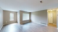 This single-floor residence offers a straightforward and functional layout finished in a neutral, contemporary palette throughout. The main living area is generously proportioned, featuring light-toned wood-style flooring that extends across the open space. Large windows fitted with vertical blinds allow natural light to filter in, brightening the room considerably. A ceiling fan overhead provides additional comfort and airflow. The adjoining kitchen is designed in a galley configuration, featuring white shaker-style cabinetry with brushed nickel hardware, grey laminate countertops, and a full suite of stainless steel appliances including a refrigerator, electric range, microwave, and dishwasher. A peninsula counter connects the kitchen to the living area, enhancing the open feel. The bedroom is a well-sized, carpeted room with its own large window offering outdoor views and ample natural light. The bathroom is finished with white ceramic tile surrounding a full soaking tub and shower combination, accented by a decorative taupe tile border, and complemented by wood-style flooring. Clean white walls and consistent neutral tones throughout the unit contribute to a cohesive, move-in ready ambiance.