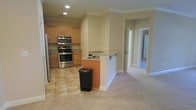 This single-floor residence offers a thoughtfully designed open-concept layout that seamlessly connects the kitchen and living areas. The kitchen is well-appointed with warm honey-toned wood cabinetry, granite countertops, and a full suite of stainless steel appliances, including a range with oven, microwave, and refrigerator. A peninsula-style breakfast bar with a built-in sink serves as both a functional workspace and a natural dividing point between the kitchen and the adjoining living space. The kitchen features tile flooring, providing a practical and easy-to-maintain surface that transitions into plush wall-to-wall carpeting in the living area. Crown molding runs throughout the space, adding a refined architectural detail to the neutral-toned walls. Recessed lighting in the kitchen ensures bright, even illumination. A hallway visible from the main living area leads to additional rooms, suggesting a well-organized floor plan with clear separation between living and private spaces. The overall ambiance is clean, neutral, and move-in ready, offering a practical yet comfortable environment suited for everyday living.