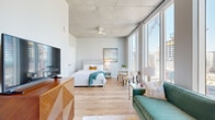 This contemporary urban studio showcases a thoughtfully designed open-concept layout that seamlessly combines living, sleeping, and dining areas under soaring exposed concrete ceilings. Light hardwood flooring flows throughout the entire space, complementing the clean, modern aesthetic.

The living area features a plush teal velvet sofa, an abstract area rug with gold accents, and elegant brass floor lamps, all positioned to take full advantage of the dramatic floor-to-ceiling windows that frame sweeping city skyline views. The open sleeping area includes a well-appointed bed with coordinating furnishings and a wall-mounted flat-screen television atop a warm wood media console.

The galley-style kitchen is outfitted with white flat-front cabinetry, a white subway tile backsplash, light stone countertops, and a full suite of stainless steel appliances including a refrigerator, range, microwave, and dishwasher. A hallway with louvered storage doors connects the kitchen to the main living space, with track lighting above adding functional illumination.

The bathroom features a frameless glass-enclosed walk-in shower with large-format neutral stone tile, offering a clean and refined finish. Throughout the space, the interplay of natural light, urban views, and modern design details creates a bright, sophisticated atmosphere well-suited to contemporary city living.