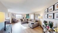 This single-floor residence is characterized by its warm, travel-inspired aesthetic and functional layout. Light hardwood flooring flows throughout the main living areas, lending a cohesive and airy feel to the space.

The expansive living room serves as the centerpiece of the home, anchored by a large sectional sofa and accented by a striking oversized world map artwork. Multiple windows allow generous natural light to fill the room, complementing the neutral wall tones and carefully curated décor including maps and framed artwork.

The dining area features a modern table and seating arrangement beneath flush-mount lighting, with a sliding glass door that opens to views of the surrounding cityscape. An entire wall is adorned with an impressive gallery of travel photographs and maps, creating a bold focal point.

Adjacent to the dining area, the kitchen is outfitted with dark cabinetry, stainless steel appliances including a gas range and refrigerator, and a secondary kitchenette with a double-basin sink and additional upper cabinetry provides extra utility.

The bedroom offers a restful retreat with plush carpeting, a ceiling fan, and a full wall of rich wood-paneled closet doors providing substantial storage. Framed artwork and soft lighting complete the comfortable ambiance throughout this well-designed home.