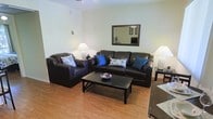 This single-floor space offers a comfortable and well-organized layout designed for easy living. The living area serves as the heart of the space, showcasing warm hardwood flooring that adds a natural richness to the room. A dark leather sofa and matching armchair provide ample seating, accented by vibrant blue and patterned throw pillows that introduce a pop of color against the neutral, light-toned walls. A rectangular dark wood coffee table anchors the seating arrangement, while a framed wall mirror reflects light and adds a sense of depth to the room. Soft ambient lighting is provided by two floor lamps positioned on either side of the seating area, contributing to a warm and inviting atmosphere. The clean, white walls enhance the brightness of the space, creating an open and airy feel throughout. The layout is efficient and thoughtfully arranged, making the most of the available space while maintaining a clear flow between areas. Overall, the space presents a tidy, functional, and welcoming environment with a classic, understated style.