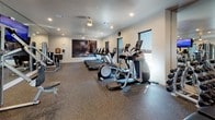 Located at 9110 Crown Springs View in Colorado Springs, Colorado, this single-story fitness center offers a comprehensive layout divided into distinct workout zones, each thoughtfully designed to support a full range of exercise activities.

The main cardio area features a row of modern treadmills positioned alongside windows that allow natural light and outdoor views, complemented by elliptical machines and upright bikes arranged on durable speckled rubber flooring. Large-scale athletic murals depicting dynamic movement adorn the walls throughout, reinforcing an energetic and motivating atmosphere. Recessed lighting and ceiling fans maintain a bright, comfortable environment.

The strength training zone is equipped with cable machines, weight benches, and a full-length mirror wall that visually expands the space. An adjacent area houses a rack of kettlebells, medicine balls, and step platforms alongside spin bikes, making it suitable for group or individual cycling and functional fitness sessions. A wall-mounted TV screen in this zone allows users to follow guided workout programming.

A smaller entry alcove provides a coat rack, storage bench, and sanitation station, offering a clean and organized transition into the workout space. Overall, the fitness center combines practical functionality with an inspiring aesthetic suited for a wide variety of training needs.