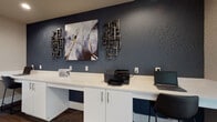 Located at 13621 Feather Sound Circle East in Clearwater, Florida, this single-floor commercial space is thoughtfully designed with a modern, professional aesthetic throughout. The standout feature is a well-equipped business center, where a continuous white built-in desk with cabinet storage runs along a striking deep-blue textured accent wall. The workspace accommodates multiple users simultaneously, outfitted with a laptop, printer, and complimentary Wi-Fi access. Decorative wall art and sculptural metallic accents lend a polished, curated character to the room. Rich, multi-toned wood-look plank flooring grounds the space with warmth and texture. A frosted glass interior window and a set of white French doors connect the business center to an adjacent common area, allowing natural light to flow through while maintaining a sense of privacy. The corridors are finished in crisp white with large-format light tile flooring, creating a bright and clean circulation path. Amenities along the hallway include clearly marked restroom facilities and a stainless steel water fountain. Recessed lighting throughout ensures consistent, even illumination across all areas. The overall ambiance is contemporary and well-maintained, reflecting a functional yet design-forward commercial environment suited for a hospitality or multi-tenant professional setting.