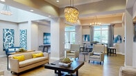 Located at 356 Lake Murray Boulevard in Irmo, South Carolina, this single-floor community space offers a generously sized, thoughtfully designed interior that blends style with functionality. The layout encompasses a living room, dining room, and office, all connected by wide, open corridors finished with wood-look flooring and neutral warm-toned walls.

The living room serves as the heart of the space, showcasing soaring ceilings with elegant crown molding, recessed lighting, and a striking sculptural chandelier as a central focal point. Multiple seating arrangements with contemporary upholstered sofas and accent chairs create inviting conversation areas, while a tiled accent wall with a wall-mounted television adds a modern touch.

The dining area transitions seamlessly from the living space, illuminated by a classic ring chandelier and framed by large windows that invite abundant natural light and outdoor views. High-top tables and chairs offer a casual yet refined dining setting.

The office is a well-appointed, multi-functional room featuring individual workstations, a large conference table with ample seating, and generous window coverage dressed with patterned curtains. Vibrant abstract artwork is displayed throughout the space, adding color and visual interest. A welcoming entry corridor with glass doors and staircase access rounds out this polished and versatile community space.