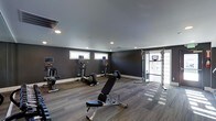 This fitness center offers a clean, contemporary workout environment designed to support a full range of exercise routines. The space is finished with dark charcoal walls and durable wood-look flooring, creating a focused and energizing atmosphere. Recessed ceiling lighting provides consistent, even illumination throughout the room, while a row of horizontal windows along one wall introduces natural light without compromising wall space. The cardio area is well-appointed with multiple upright elliptical machines and additional cardiovascular equipment, giving users ample options for aerobic training. A sturdy adjustable workout bench occupies the center of the floor, ideal for strength and conditioning exercises. Along the far wall, a multi-functional cable and pulley machine provides versatile resistance training capabilities. A glass-panel door offers easy access to the space while contributing to an open, airy feel. The layout is thoughtfully organized to allow comfortable movement between stations, making the space functional for both individual workouts and simultaneous use by multiple users. Overall, the fitness center presents a polished, professional aesthetic that balances form and function.