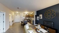 This single-floor space showcases a well-designed open-concept layout that integrates the kitchen, dining, and living areas into one cohesive environment. The kitchen is equipped with stainless steel appliances, including a refrigerator, oven, and dishwasher, complemented by dark wood cabinetry that extends to the upper walls for generous storage. A kitchen island with seating provides a casual dining option and serves as a natural dividing point between the kitchen and living areas. The flooring throughout is a warm-toned wood-look material that adds continuity and visual warmth across the space. The living area is anchored by a neutral sofa and accented by a bold navy feature wall adorned with gold geometric décor and framed artwork, creating a striking focal point. A flat-screen television is mounted on a media console nearby. Recessed and flush-mount lighting fixtures illuminate the space evenly, while white walls and smooth ceilings contribute to a bright, clean aesthetic. A white paneled entry door is visible, offering a clear sense of the unit's layout and flow from entry to living space.