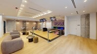 Located in the heart of downtown Miami at 55 NE 2nd St, this single-floor amenity space is a thoughtfully designed entertainment destination offering a diverse range of social and recreational experiences. The centerpiece of the space is a pair of full-length bowling lanes, dramatically accented by a glowing neon "BOWL" sign and overhead flat-screen monitors, bringing a lively, energetic atmosphere to the room. Light wood flooring runs throughout, complementing the clean white walls and recessed lighting that maintain a modern, polished aesthetic. Adjacent to the bowling area, a generous lounge features plush rounded sofas and layered coffee tables, creating an inviting space for relaxation. A striking wet bar serves as a social hub, finished with a dark stone countertop, bold red tile detailing, and warm yellow barstools beneath a row of globe pendant lights. Floor-to-ceiling glass walls frame sweeping city views, flooding the space with natural light. A glass-enclosed room provides a quieter retreat, while a separate dining nook with vertical wood slat accent walls and bistro-style high-top tables adds a casual yet refined dining option. The overall design blends upscale finishes with vibrant entertainment elements, making it an exceptional communal destination.