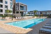 Located at 100 Zoo Ln in Santa Ana, California, this outdoor amenity space presents a striking resort-style pool deck designed for relaxation and leisure. The centerpiece is a generously sized swimming pool with crystal-clear blue water, accessible via wide entry steps and framed by a spacious paver deck finished in warm neutral tones. Rows of contemporary lounge chairs are arranged along the poolside, offering ample space for sunbathing beneath the Southern California sky. Lush tropical landscaping, including tall palm trees, ornamental grasses, and vibrant plantings, adds a verdant, resort-inspired atmosphere throughout the space. Pergola-style shade structures provide sheltered seating areas, complemented by additional lounge furnishings including wicker pod chairs. The pool deck is enclosed within a courtyard formed by modern multi-story residential buildings featuring clean white and earth-toned facades, large windows, and private balconies with glass railings. The architectural design creates a semi-private, open-air retreat that balances urban sophistication with a relaxed, tropical ambiance. The overall environment is bright, airy, and thoughtfully landscaped, offering residents an inviting outdoor sanctuary in the heart of Santa Ana.