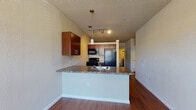 This single-floor residence offers a well-organized, open layout designed for comfortable everyday living. The kitchen serves as the heart of the home, featuring warm cherry-toned cabinetry, dark granite-style countertops, and a full suite of black appliances including a refrigerator, electric range, and built-in microwave. A peninsula with an integrated sink extends into the living area, providing additional prep space and a natural division between the kitchen and the open living zone. Track lighting and a pendant light above the peninsula ensure the space is well illuminated throughout.

The living and dining area benefits from rich hardwood-style flooring, neutral-toned walls, and a large window flanked by a front entry door, allowing natural light to fill the space. A ceiling fan adds both function and comfort to this area.

The bedroom is carpeted for added warmth and comfort, featuring a thermostat on the wall and direct access to a hallway leading to additional spaces.

The bathroom includes a granite-style vanity countertop with an undermount sink, offering practical storage and countertop space. A hallway off the bathroom reveals a stacked in-unit washer and dryer, adding notable convenience. Throughout the residence, textured walls and consistent neutral finishes create a cohesive and functional living environment.
