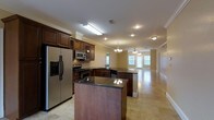 This single-floor space is defined by its open-concept layout, seamlessly connecting the kitchen and living areas to create a generous, flowing interior. The kitchen is a standout feature, showcasing rich dark wood cabinetry that extends to the upper walls, complemented by dark granite countertops that add a touch of elegance. A central island provides additional prep and gathering space. Stainless steel appliances, including a French-door refrigerator with an ice and water dispenser, a gas range with a built-in microwave above, and a sink integrated into the extended countertop peninsula, make this a highly functional cooking environment. The light travertine tile flooring runs throughout, lending a warm, cohesive feel to the space. Crown molding adorns the ceilings, adding a refined architectural detail. The living area benefits from abundant natural light streaming through large windows and glass doors, creating a bright and welcoming ambiance. Ceiling fans and a chandelier provide both practical lighting and decorative character. The overall design balances classic finishes with an open, airy atmosphere well-suited for both everyday living and entertaining.