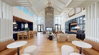 Located at 1009 Reagan Road in Frederick, Maryland, this single-floor community clubhouse showcases an impressive blend of modern design and warm, inviting materials. The centerpiece of the space is a grand great room with soaring cathedral ceilings framed by white-painted wood beam detailing and a monumental floor-to-ceiling stacked stone fireplace that commands the room. Expansive floor-to-ceiling windows and multi-panel black-framed glass doors flood the interior with abundant natural light and offer views of the surrounding outdoor areas. Wide-plank light oak hardwood flooring runs throughout, tying together the open-plan layout seamlessly. The great room accommodates multiple lounge seating arrangements, including plush sofas, curved accent chairs, and a communal dining area, providing versatile spaces for gathering and relaxation. A dedicated media wall features custom navy cabinetry with glass-front displays and warm wood accents flanking a mounted television. A separate kitchen area is finished with deep navy shiplap walls, a natural wood-beam ceiling, sleek slate-toned cabinetry, gold hardware, and striking veined stone countertops with backsplash. A glass-partitioned entryway with black steel framing creates an elegant transition into the main hall. Built-in bench seating with a white slatted privacy screen adds a cozy nook within the social space.