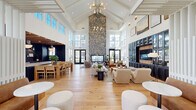 Located at 1009 Reagan Road in Frederick, Maryland, this single-floor community clubhouse showcases an impressive blend of modern design and warm, inviting materials. The centerpiece of the space is a grand great room with soaring cathedral ceilings framed by white-painted wood beam detailing and a monumental floor-to-ceiling stacked stone fireplace that commands the room. Expansive floor-to-ceiling windows and multi-panel black-framed glass doors flood the interior with abundant natural light and offer views of the surrounding outdoor areas. Wide-plank light oak hardwood flooring runs throughout, tying together the open-plan layout seamlessly. The great room accommodates multiple lounge seating arrangements, including plush sofas, curved accent chairs, and a communal dining area, providing versatile spaces for gathering and relaxation. A dedicated media wall features custom navy cabinetry with glass-front displays and warm wood accents flanking a mounted television. A separate kitchen area is finished with deep navy shiplap walls, a natural wood-beam ceiling, sleek slate-toned cabinetry, gold hardware, and striking veined stone countertops with backsplash. A glass-partitioned entryway with black steel framing creates an elegant transition into the main hall. Built-in bench seating with a white slatted privacy screen adds a cozy nook within the social space.